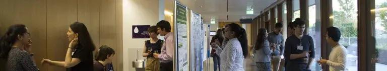 Photo of symposium attendees standing in groups chatting, with drinks and looking at posters