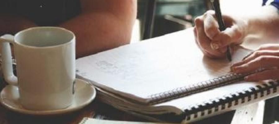 Photo of notepads open on a table, witha a pen, hands writing and the shoulder of someone sitting at the table