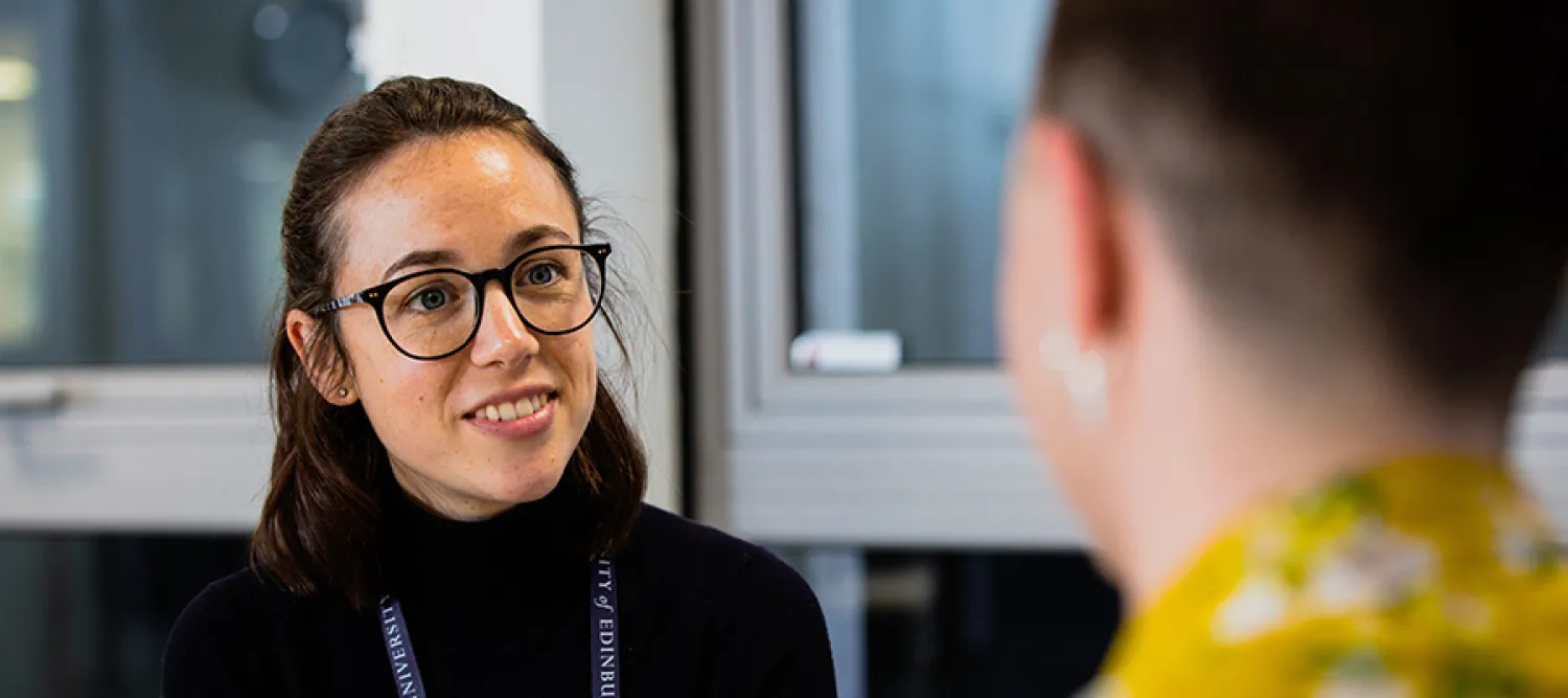 Photo of a woman wearing glasses who smiles, whilst listening to a student who sits opposite, with their back to the camera