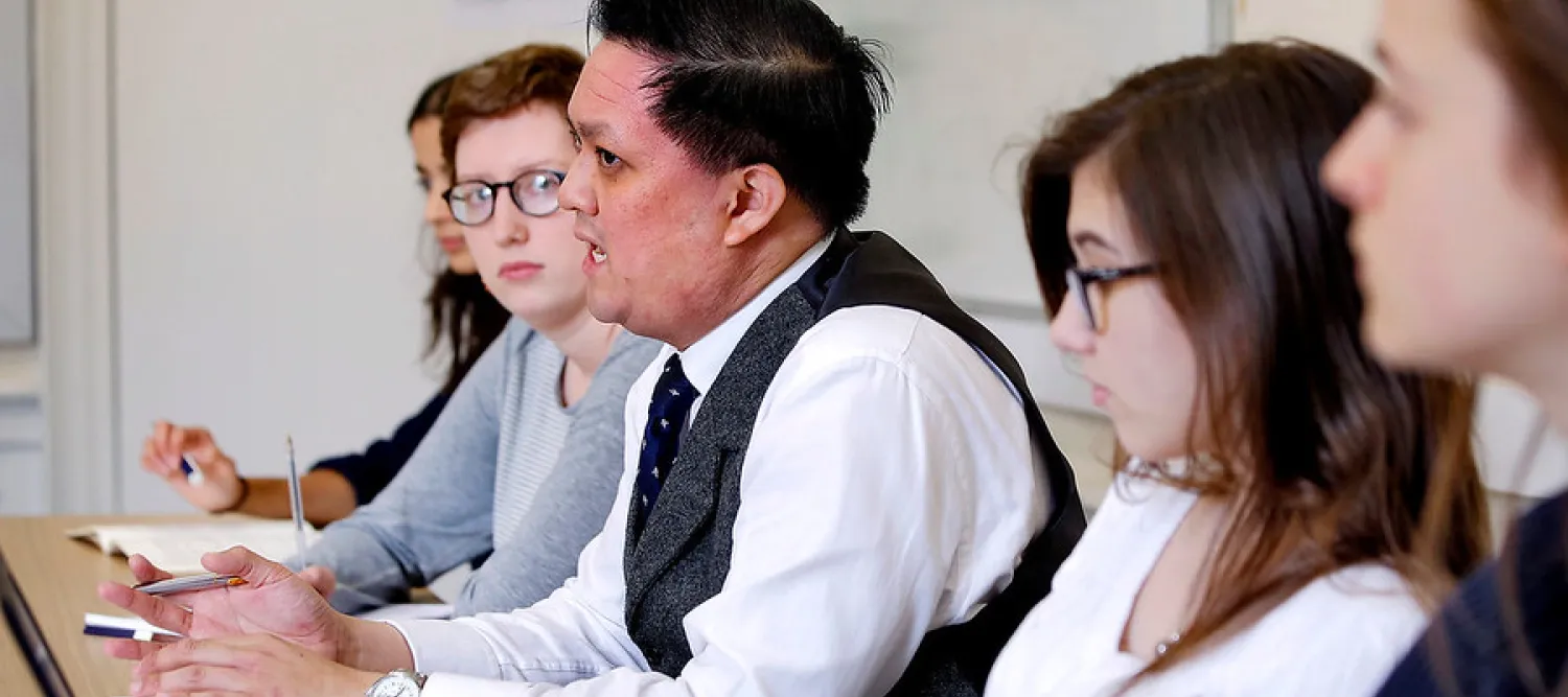 A group of people sit next to each other at a long table, whilst the middle person speaks 