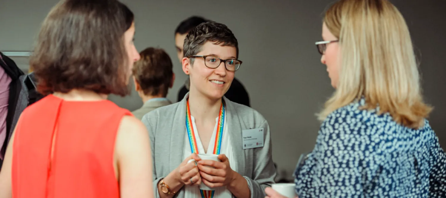 Photo of 3 people standing chatting, one wears a university rainbow lanyard