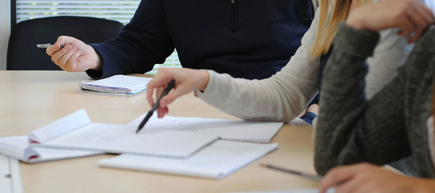 Photo of people sitting around a table, although you can't see their head/faces, on the table are papers and people hold pens hovered over them