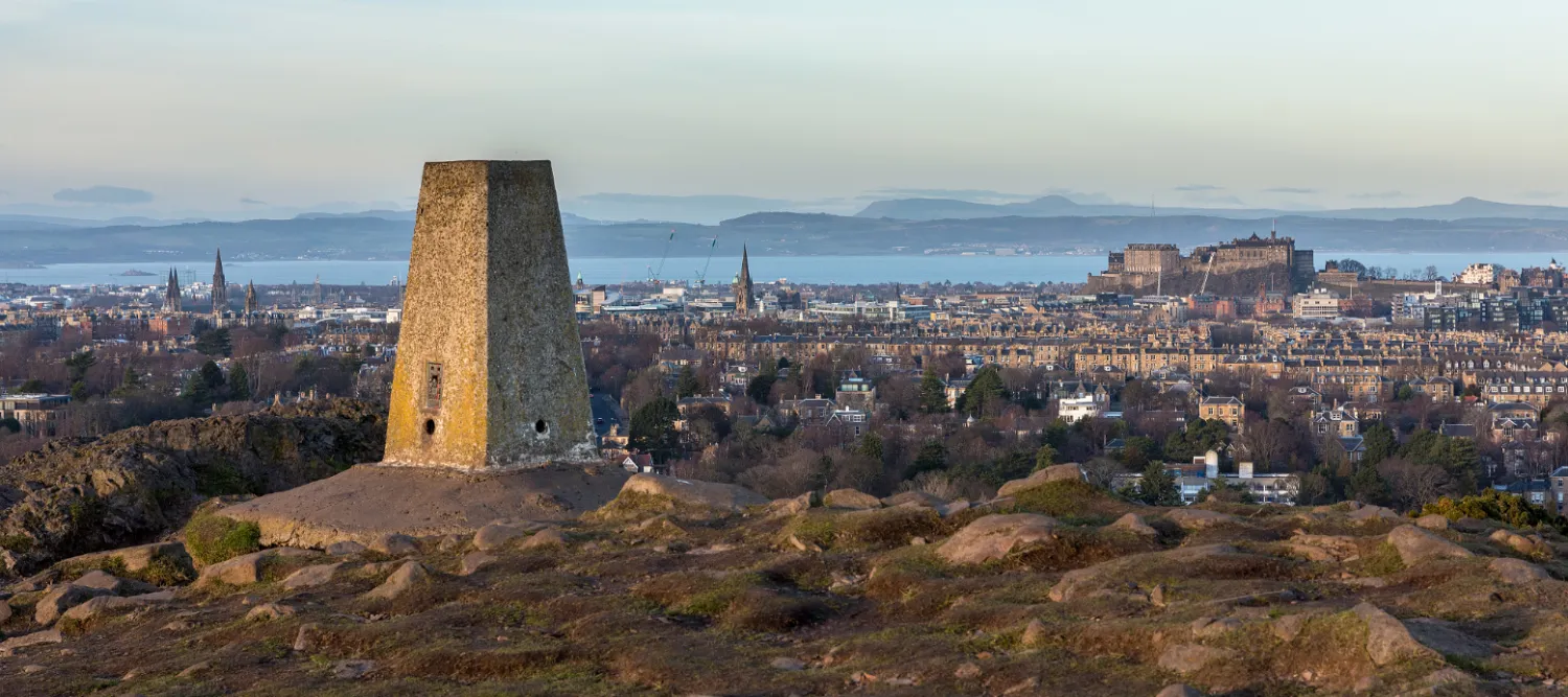 View from the top of Blackford Hill. There is a white trig point in the foreground with views over to Edinburgh Castle and Fife in the background.