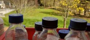 A number of jars with chemicals on a window sill with the view of trees outside of the window.