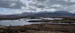 Photo of a landscape with a grassy moore and a body of water in the foreground and hills in the background, the sky above is mostly cloudy