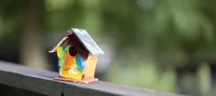 A small colourfully painted birdbox on a fence in front of trees
