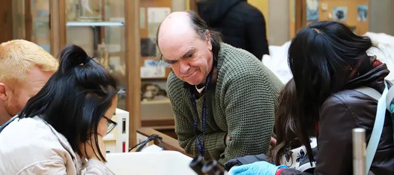 A member of staff leans over a workbench with students nearby also looking at workbech 