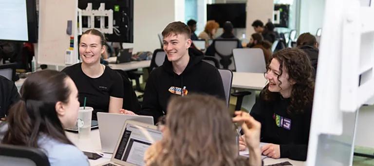 Students sit at desks in a cluster using laptops and chatting