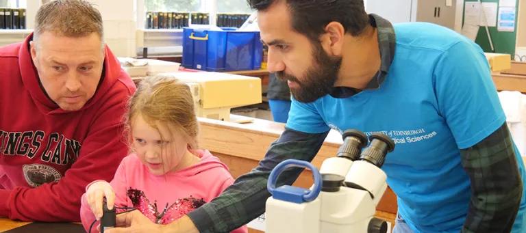 Photo of a School researcher wearing a blue School t-shirt and showing a parent and child images from a microscope