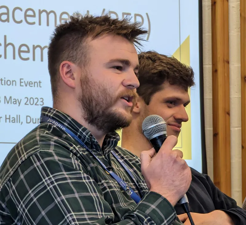 Two people on stage in front of a screen, one holds and is talking into a microphone