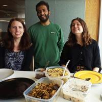 Three group members stand in front of a table with food in containers and plates on it.