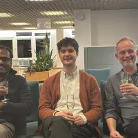 Thom sits between JP and Kevin Hrdwick in a seated area, they each have a glass of champagne