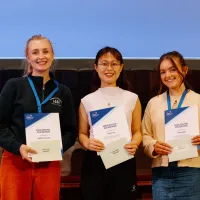 Three people holding certificates at an award event, wearing blue lanyards, with a screen and wooden paneling behind them.