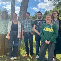 Six people stand infront of trees and bushes on a sunny day