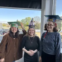 Dr Zishan Fu, Dr Tea Reinert, and Dr Laura Cooper stand in front of a window wearing crafted paper hats, celebrating passing their vivas