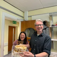 Two people in a room, one holding a birthday cake with toasted meringue and a “Happy Birthday” topper, with shelves and a microwave in the background.