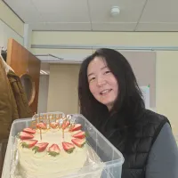 Person indoors holding a round birthday cake topped with strawberries and gold “Happy Birthday” topper, wearing a black vest over a grey shirt, with a coat hanging on a door in the background.