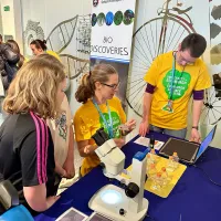 Photo of three group members in yellow t-shirts holding props and standing in front of a table of props for Edinburgh Science Festival