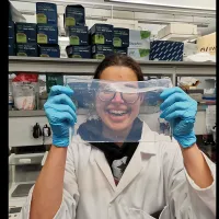 Photo of Alex in the lab, in a lab coat and blue gloves, holding up a piece of transparent material in front of her
