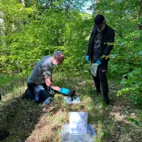 Two fieldworkers in a clearing in a wooded area use a camera on the ground to take photos