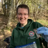 Photo of a fieldworker wearing a green hoodie and blue gloves holding up a fieldmouse for the camera