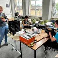 YuPhoto of three research group members in a classroom, with desks and chairs working on black boxes used in their work