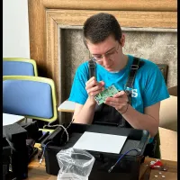 Photo of a group member sitting at a table working on a circuit board with a large black box sitting on the table in front of them