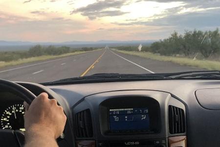 Looking out from the front seat of a car, you can see the dashboard and a long straight road ahead.