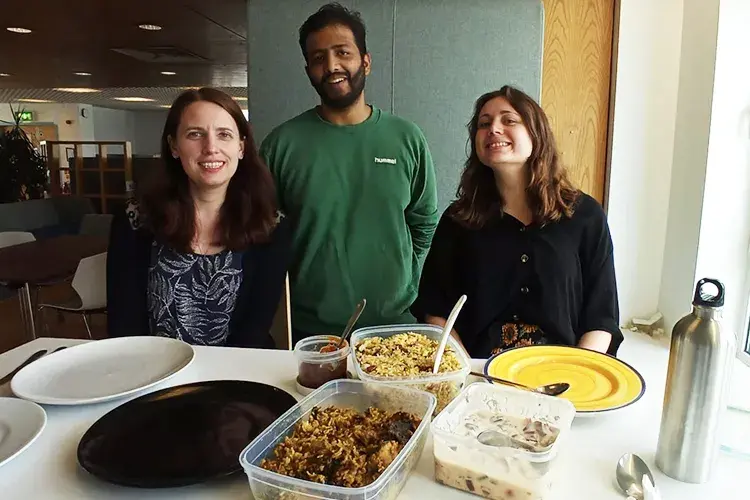 Three group members stand in front of a table with food in containers and plates on it.