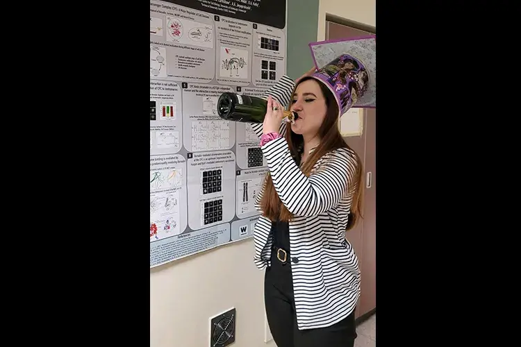 Paula stands in front of a research poster wearing a big hat and swigging from a bottle of champagne