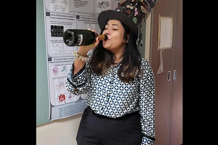 Pragya stands in front of a research poster, wearing a large hat and sigging a bottle of champagne