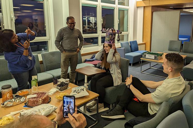 Paula tries on a large, colourful hat whilt surrounded by group members in a seated area