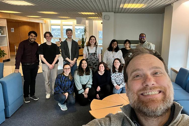 Group photo with lab members in the background and someone standing in front of the camera taking the photo in a selfie style