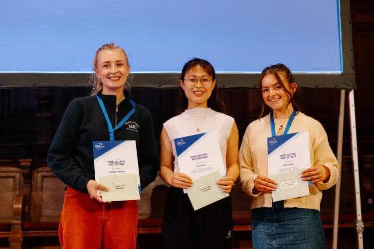 Three people holding certificates at an award event, wearing blue lanyards, with a screen and wooden paneling behind them.