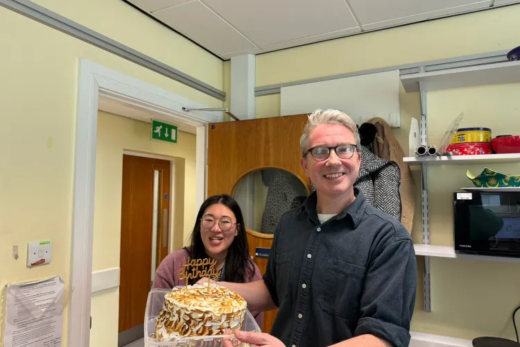 Two people in a room, one holding a birthday cake with toasted meringue and a “Happy Birthday” topper, with shelves and a microwave in the background.