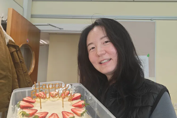 Person indoors holding a round birthday cake topped with strawberries and gold “Happy Birthday” topper, wearing a black vest over a grey shirt, with a coat hanging on a door in the background.