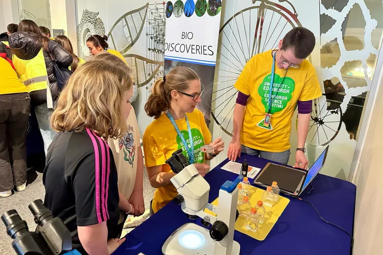 Photo of three group members in yellow t-shirts holding props and standing in front of a table of props for Edinburgh Science Festival