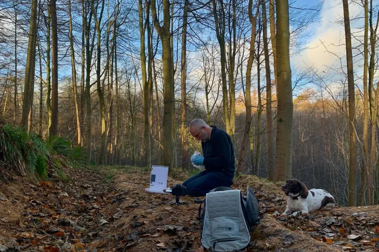 Photo of a fieldworker sitting working in a clearing in a wooded area, a brown and white spaniel sitting nearby
