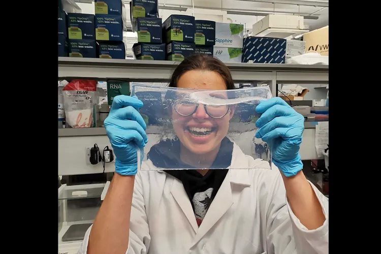 Photo of Alex in the lab, in a lab coat and blue gloves, holding up a piece of transparent material in front of her
