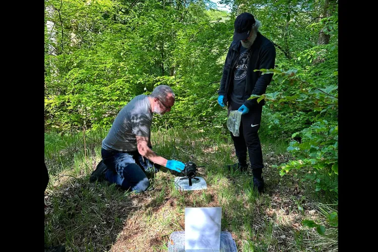 Two fieldworkers in a clearing in a wooded area use a camera on the ground to take photos