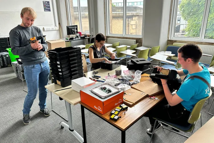 YuPhoto of three research group members in a classroom, with desks and chairs working on black boxes used in their work