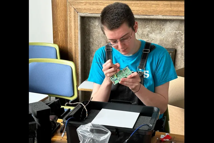 Photo of a group member sitting at a table working on a circuit board with a large black box sitting on the table in front of them