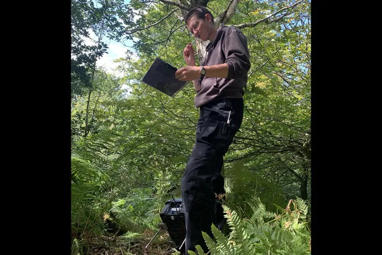 Photo of a fieldworker standing in a wooded area with a clipboard and pen