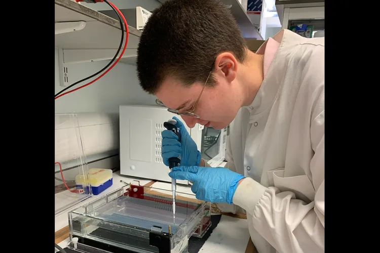 Photo of a research group member in the lab, wearing a white coat and blue gloves, using a pippet