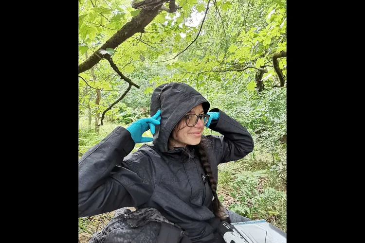Photo of Alex ina hooded jacket wearing blue gloves holdoing up her hood, she's in a wooded are and has a bag and datasheets in a folder