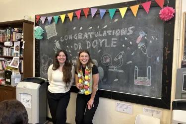 Photo of Rose Doyle and Maddie Moule in front of chalkboard that reads "Congratulations Dr Rose Doyle"