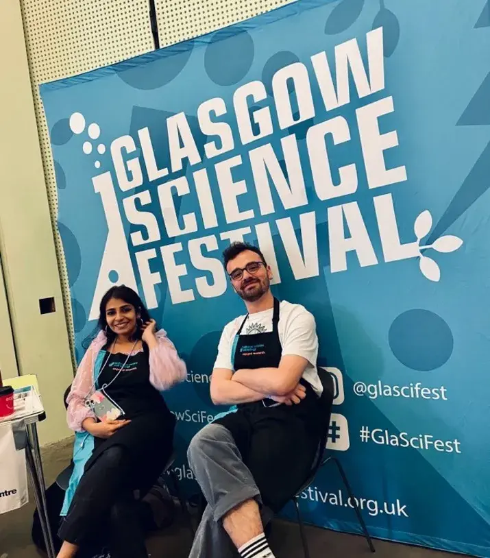 Two lab group members sit wearing aprons in front of a Glasgow Scieence Festival poster