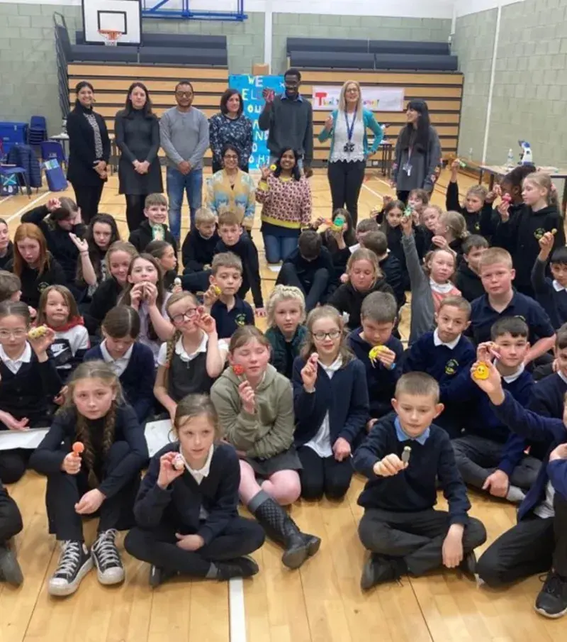 A group of school children sit on a gym floor showing what they created during a workshop, researchers stand in the background