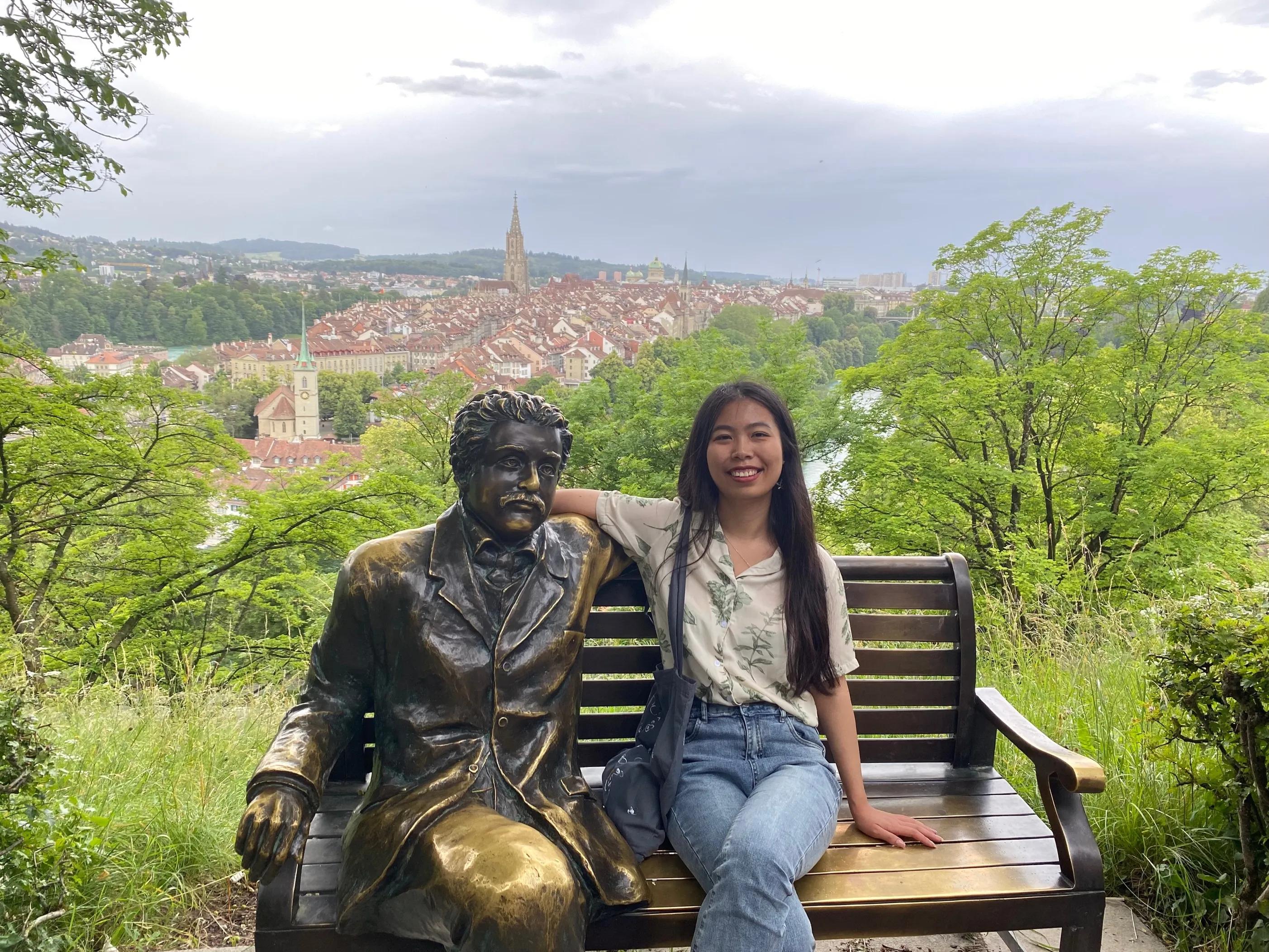Photo of Haomio Cheng sitting on a bench beside a bronze statue with a city scape behind her