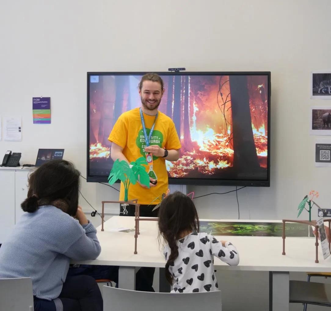Photograph of a young man wearing a yellow Edinburgh Science Festival T-shirt talking to workshop participants in a classroom with a TV screen behind showing a forest fire